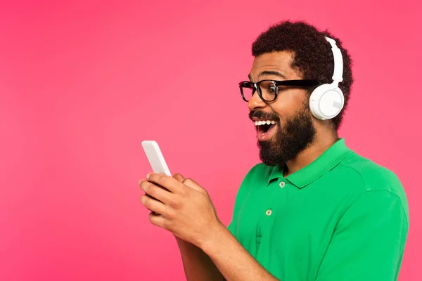 Aufgeregter afrikanisch-amerikanischer Mann mit Brille und drahtlosen Kopfhörern, Smartphone isoliert auf rosa — Stockfoto