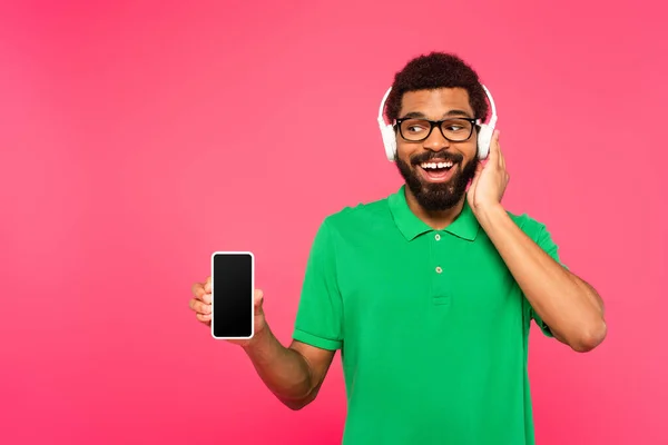 Hombre afroamericano en gafas y auriculares inalámbricos sosteniendo teléfono inteligente con pantalla en blanco aislado en rosa - foto de stock