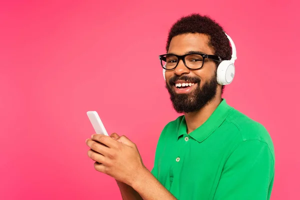 Hombre afroamericano en gafas y auriculares inalámbricos mensajes de texto en el teléfono inteligente aislado en rosa - foto de stock