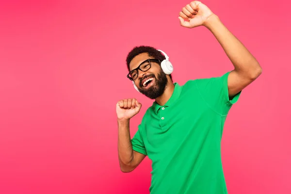 Збуджений афроамериканський чоловік в окулярах і бездротових навушниках слухає музику на рожевому — стокове фото
