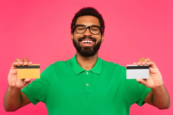 Fröhlicher afrikanisch-amerikanischer Mann mit Brille und Kreditkarten isoliert auf rosa — Stockfoto