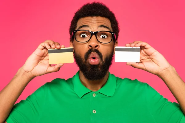 Shocked african american man in glasses holding credit cards isolated on pink — Stock Photo