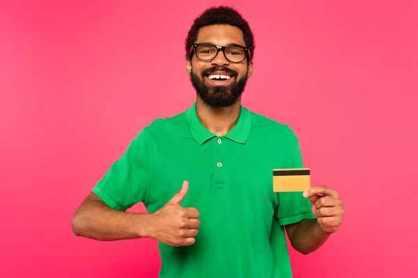 Heureux homme afro-américain dans des lunettes tenant carte de crédit et montrant pouce vers le haut isolé sur rose — Photo de stock