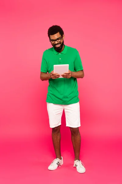 Pleine longueur de heureux homme afro-américain en lunettes, short et polo vert en utilisant une tablette numérique sur rose — Photo de stock