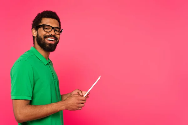 Homem americano africano alegre em óculos e camisa polo verde usando tablet digital isolado em rosa — Fotografia de Stock