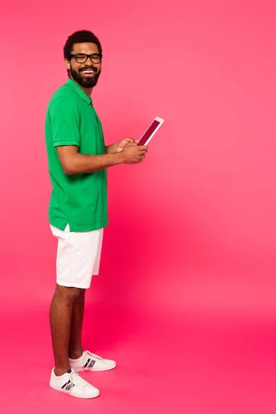 Full length of smiling african american man in glasses, shorts and green polo shirt using digital tablet on pink — Stock Photo