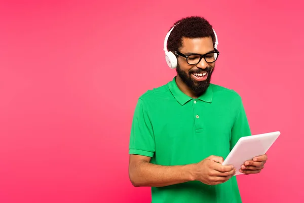 Homem americano africano alegre em óculos e fones de ouvido usando tablet digital isolado em rosa — Fotografia de Stock