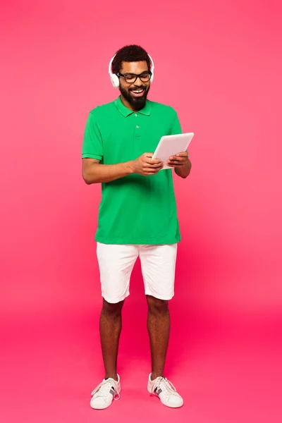 Pleine longueur de l'homme afro-américain joyeux dans des lunettes et des écouteurs en utilisant une tablette numérique sur rose — Photo de stock