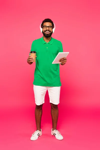 Piena lunghezza di allegro uomo afroamericano in occhiali e cuffie in possesso di tazza di carta e tablet digitale su rosa — Foto stock