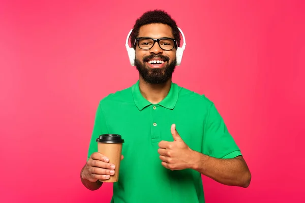 Homem americano africano alegre em óculos e fones de ouvido segurando copo de papel e mostrando polegar isolado em rosa — Fotografia de Stock