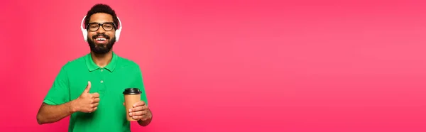 Веселий афроамериканський чоловік в окулярах і навушниках тримає паперову чашку і показує великий палець вгору ізольовано на рожевому, банер — стокове фото
