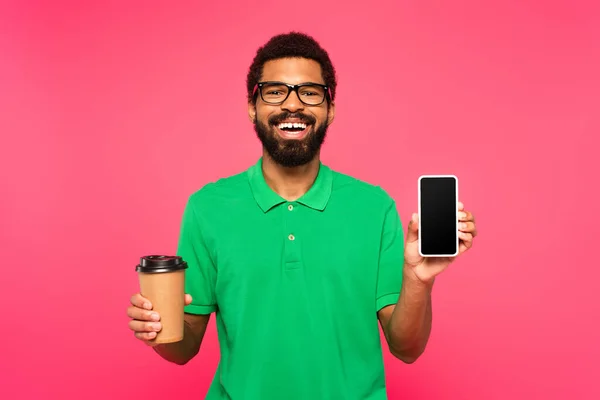 Homem americano africano feliz em óculos e camisa polo verde segurando copo de papel e smartphone com tela em branco isolado em rosa — Fotografia de Stock