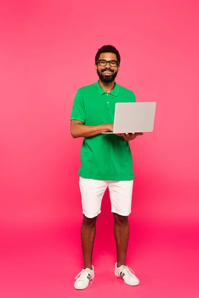 Pleine longueur de heureux homme afro-américain en lunettes, short et polo vert tenant ordinateur portable sur rose — Photo de stock