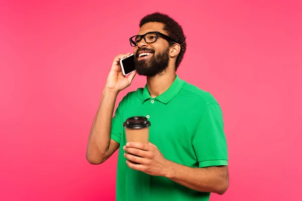 Hombre afroamericano en gafas y polo verde hablando en el teléfono inteligente mientras sostiene taza de papel aislado en rosa - foto de stock