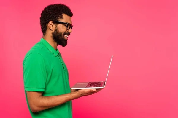 Vista lateral do homem americano africano feliz em óculos e camisa de pólo verde segurando laptop isolado em rosa — Fotografia de Stock