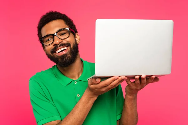 Homme afro-américain positif dans des lunettes et un polo vert tenant ordinateur portable isolé sur rose — Photo de stock