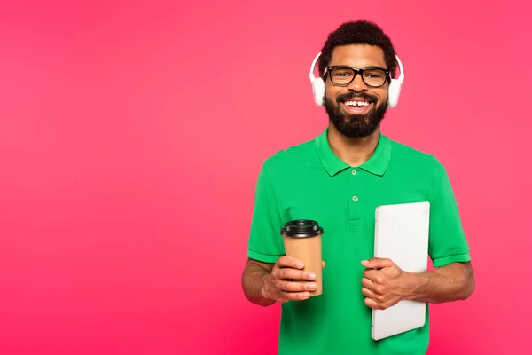 Felice uomo africano americano in occhiali e polo verde con tazza di carta con computer portatile isolato su rosa — Foto stock
