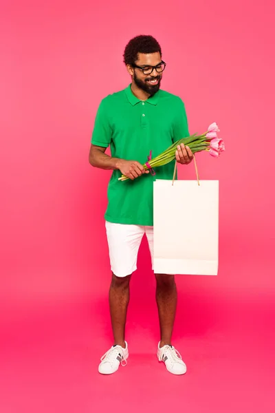 Щасливий афроамериканський чоловік в окулярах і зеленій сорочці поло, що тримає тюльпани і паперовий мішок ізольовані на рожевому — стокове фото