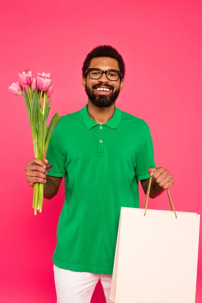 Happy african american man in glasses and green polo shirt holding tulips and shopping bag isolated on pink — Stock Photo