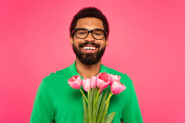 Homem americano africano alegre em óculos e camisa de pólo verde segurando tulipas isoladas em rosa — Fotografia de Stock