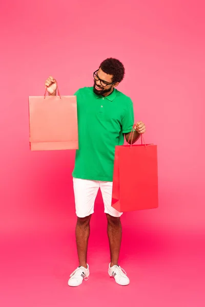 Piena lunghezza di felice uomo afroamericano in occhiali e polo verde che tiene borse della spesa su rosa — Foto stock
