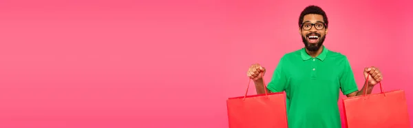 Homme afro-américain étonné dans des lunettes et un polo vert tenant des sacs à provisions isolés sur rose, bannière — Photo de stock