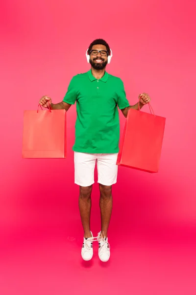 Comprimento total de homem americano africano feliz em óculos e fones de ouvido segurando sacos de compras em rosa — Fotografia de Stock