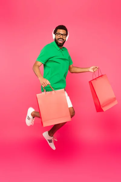 Comprimento total de homem americano africano animado em óculos e fones de ouvido segurando sacos de compras em rosa — Fotografia de Stock