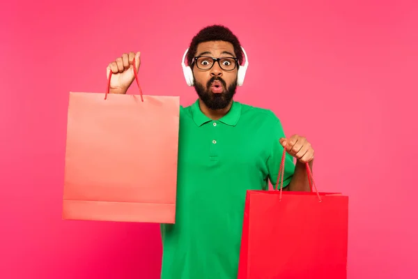 Chocado afro-americano homem em óculos e fones de ouvido segurando sacos de compras em rosa — Fotografia de Stock