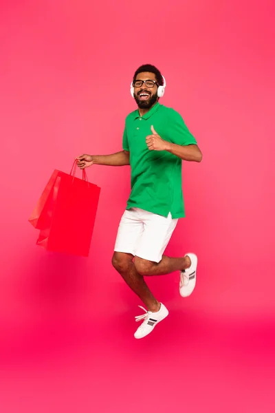 Pleine longueur de l'homme afro-américain dans des écouteurs tenant des sacs à provisions et montrant pouce vers le haut sur rose — Photo de stock