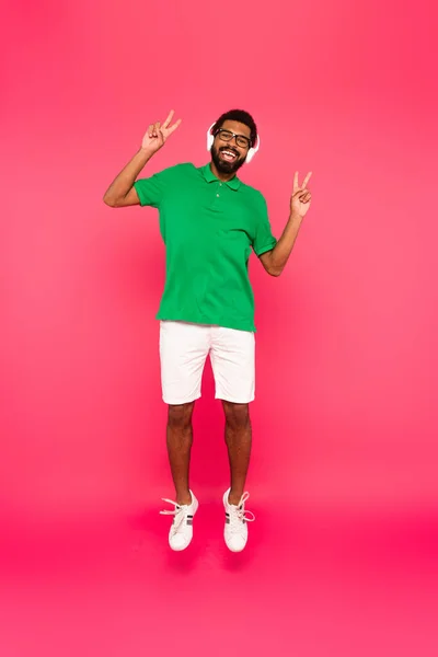 Comprimento total de homem americano africano feliz em fones de ouvido mostrando sinal de paz e pulando em rosa — Fotografia de Stock