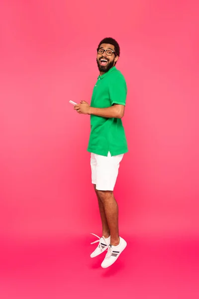 Pleine longueur de l'homme afro-américain étonné en lunettes, short et polo vert tenant smartphone sur rose — Photo de stock