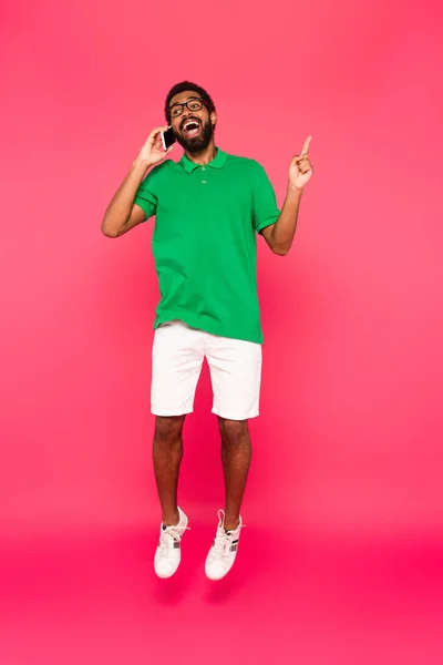 Longitud completa de hombre afroamericano emocionado en gafas saltando mientras habla en el teléfono inteligente en rosa - foto de stock