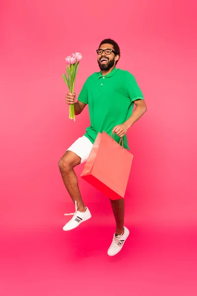 Volle Länge des glücklichen afrikanisch-amerikanischen Mannes in Brille mit Einkaufstasche und Tulpenstrauß auf rosa — Stockfoto