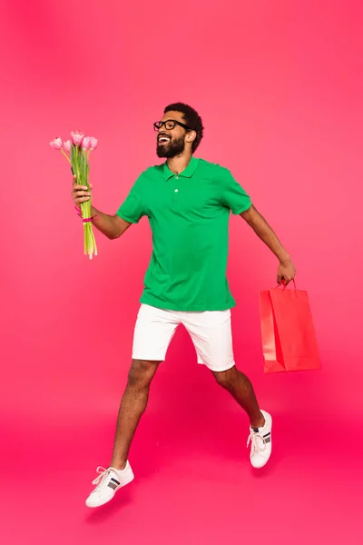 Volle Länge des glücklichen afrikanisch-amerikanischen Mannes in Brille und grünem Poloshirt mit Einkaufstasche und Tulpenstrauß auf rosa — Stockfoto