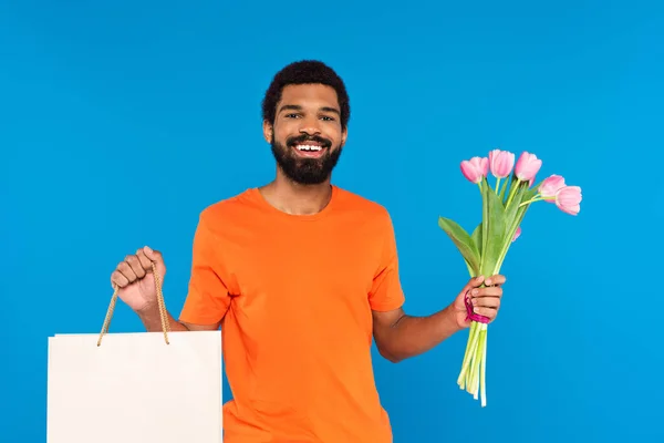 Hombre afroamericano feliz sosteniendo tulipanes rosados y bolsa aislada en azul - foto de stock