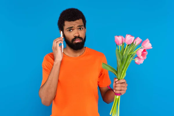 Hombre afroamericano sosteniendo tulipanes rosados y hablando en teléfono inteligente aislado en azul - foto de stock