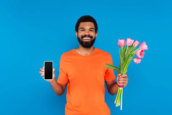 Felice uomo africano americano che tiene tulipani rosa e smartphone isolato su blu — Foto stock