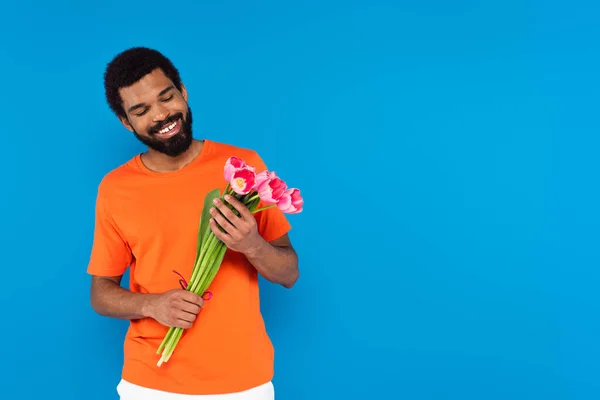 Hombre afroamericano feliz sosteniendo ramo de tulipanes rosados aislados en azul - foto de stock