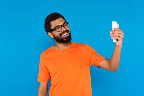 Бородатий афроамериканський чоловік в окулярах посміхається і приймає селфі ізольовано на синьому — стокове фото