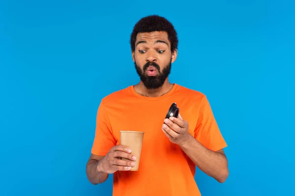 Удивленный африканский американец смотрит на кофе, чтобы пойти в бумажной чашке изолированы на синий — стоковое фото