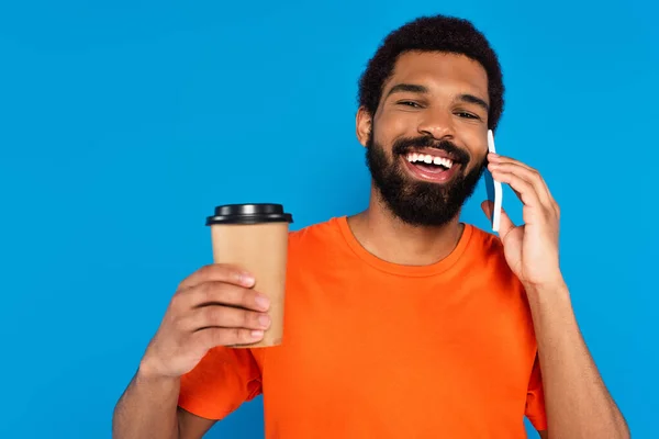 Hombre afroamericano feliz sosteniendo taza de papel y hablando en el teléfono inteligente aislado en azul - foto de stock
