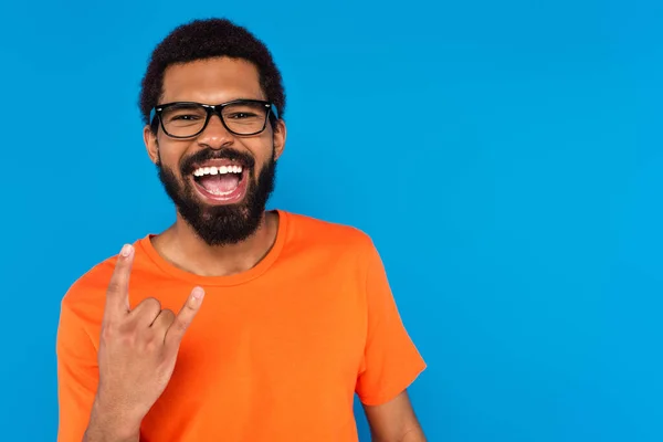 Africano americano hombre en gafas sonriendo y mostrando roca signo aislado en azul - foto de stock