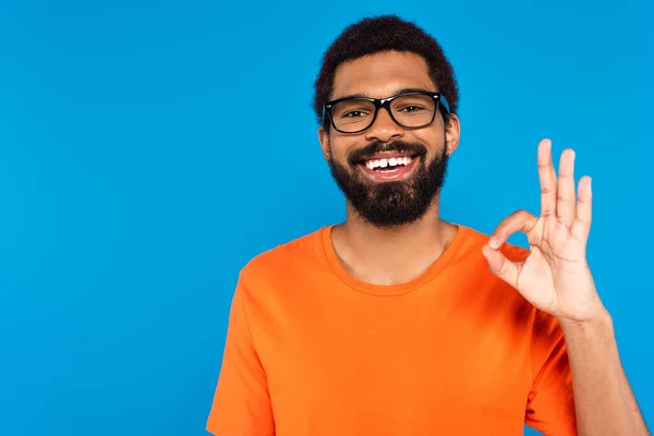 Щасливий афроамериканський чоловік в окулярах, показуючи знак ОК ізольовано на синьому — стокове фото