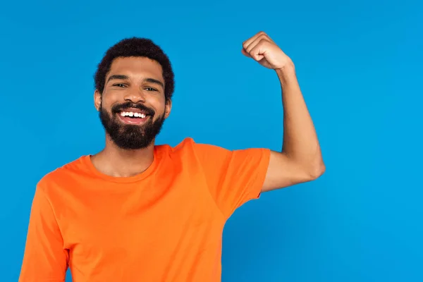 Barbudo afroamericano hombre sonriendo y mostrando músculo aislado en azul - foto de stock