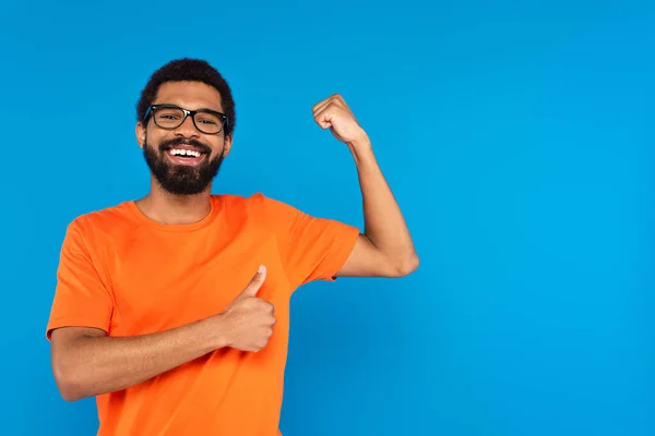 Hombre afroamericano feliz en gafas sonriendo, mostrando el músculo y el pulgar hacia arriba aislado en azul - foto de stock