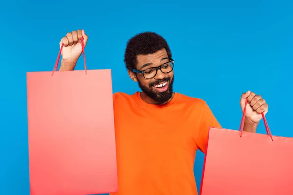 Felice uomo africano americano in occhiali che tiene borse della spesa isolate sul blu — Foto stock