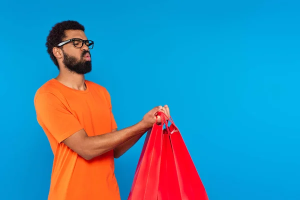 African american man in glasses holding shopping bags and pouting lips isolated on blue — Stock Photo