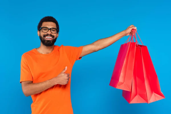 Щасливий афроамериканський чоловік в окулярах тримає сумки для покупок і показує великий палець вгору ізольовано на синьому — стокове фото