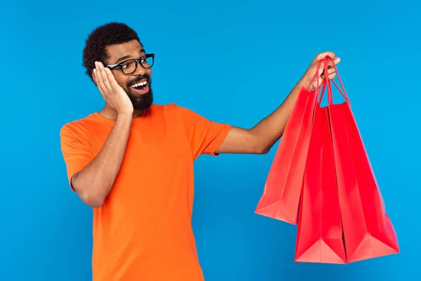 Homem americano africano espantado em óculos segurando sacos de compras isolados em azul — Fotografia de Stock